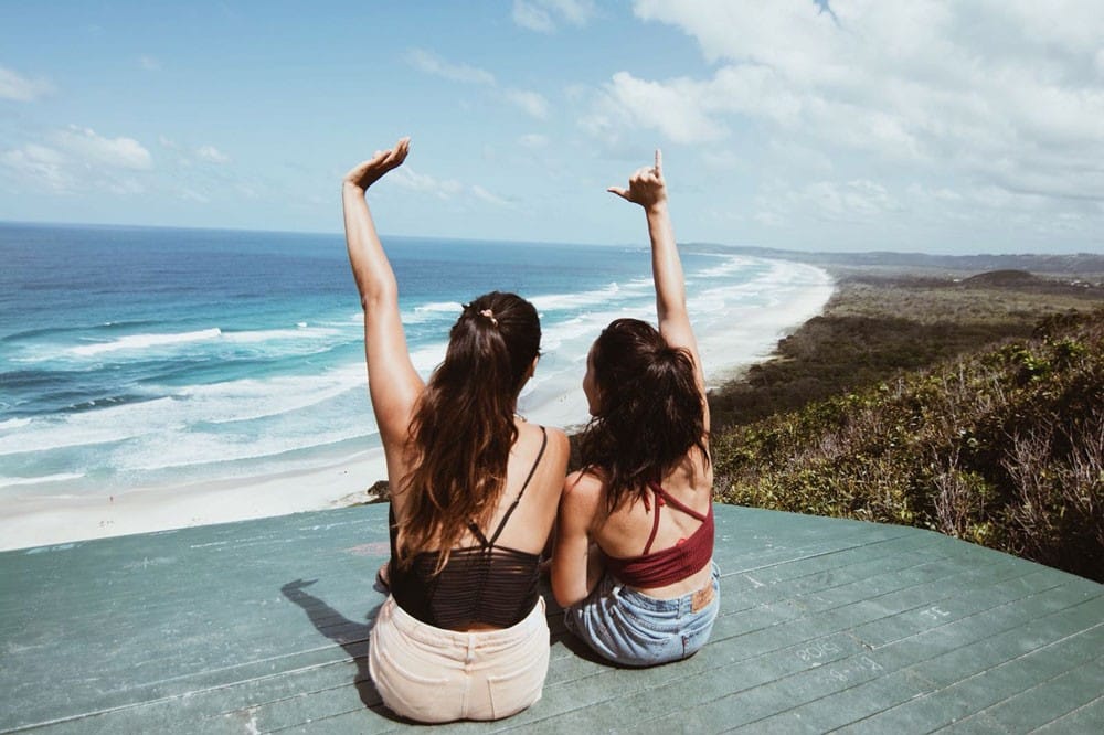 dos chicas de espaldas sentadas en un mirador de Byron Bay con vistas a la playa